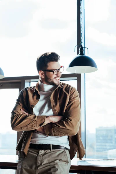 Niedrigwinkelblick eines IT-Mitarbeiters mit verschränkten Armen, der lächelt und in der Nähe von Fenstern wegschaut — Stockfoto