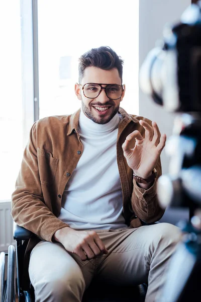 Foco seletivo de blogueiro com deficiência sorrindo e mostrando sinal ok na cadeira de rodas — Fotografia de Stock