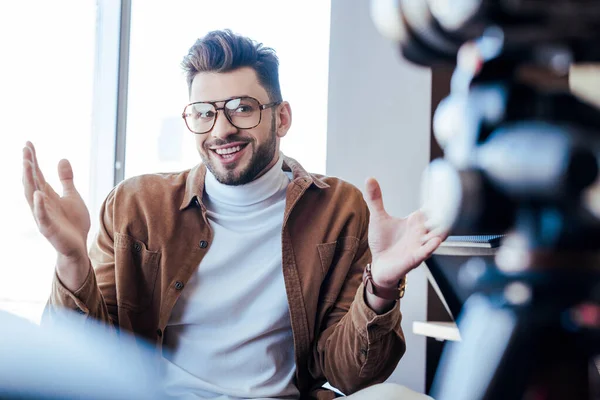 Focus selettivo del blogger in occhiali sorridenti e gesticolanti vicino alle finestre — Foto stock