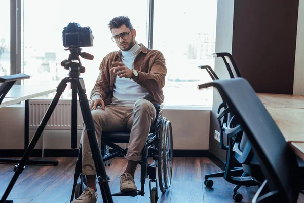 Selective focus of disabled blogger on wheelchair pointing with finger in front of digital camera near windows — Stock Photo
