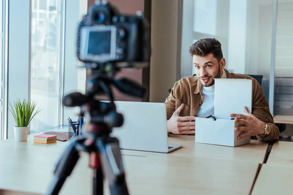 Selective focus of blogger pointing at box near laptop at table in front of digital camera — Stock Photo