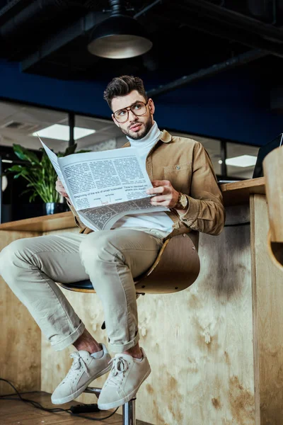 Vista de bajo ángulo del trabajador de TI con periódico mirando hacia otro lado en la silla en el espacio de coworking - foto de stock