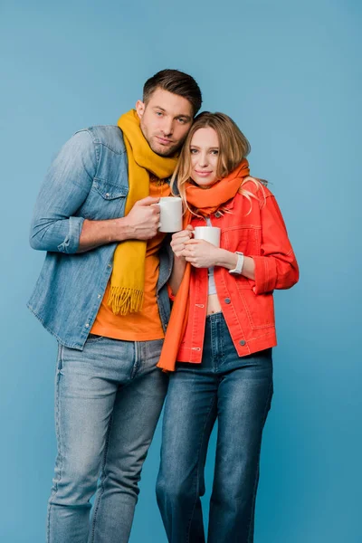 Ill couple in scarves holding cups with hot drinks isolated on blue — Stock Photo