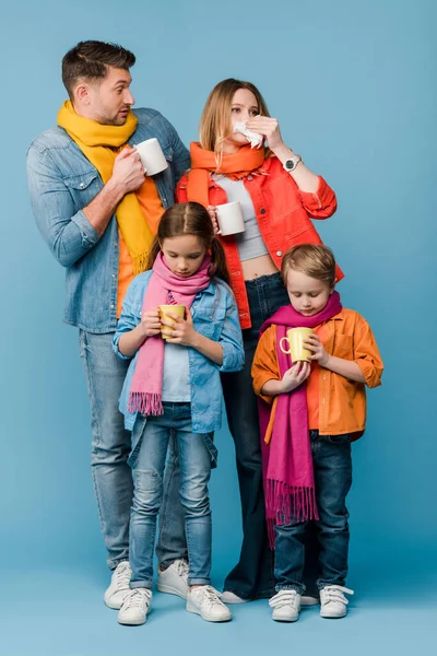 Triste famille malade avec des enfants dans des foulards tenant des tasses avec des boissons chaudes sur bleu — Photo de stock