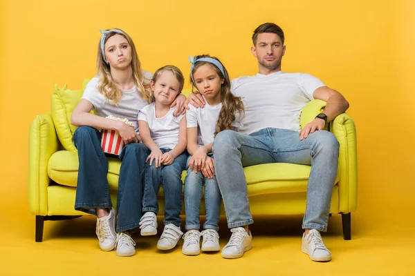 Família triste assistindo filme no sofá com balde de pipoca no amarelo — Fotografia de Stock