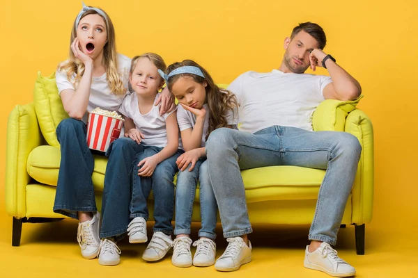 Bored parents and kids watching movie on sofa with popcorn bucket on yellow — Stock Photo