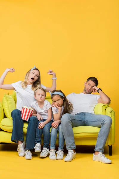 Скучающая семья зевает и смотрит кино на диване с ведром попкорна на желтом — стоковое фото