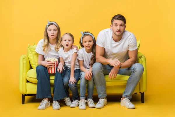 Surprised family watching movie on sofa with popcorn bucket on yellow — Stock Photo