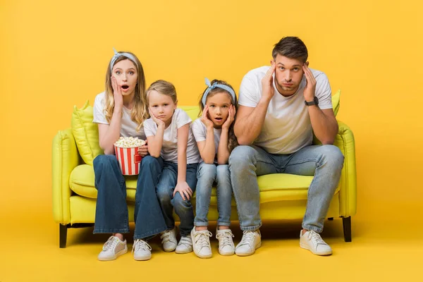 Sorprendidos padres y niños viendo la película en el sofá con palomitas de maíz cubo en amarillo - foto de stock