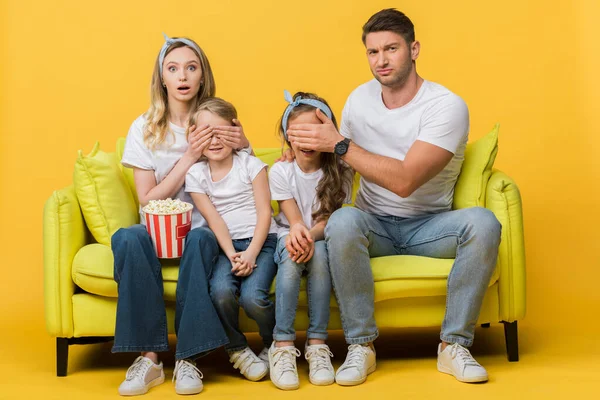 Confused parents closing eyes to kids while watching movie on sofa with popcorn bucket on yellow — Stock Photo