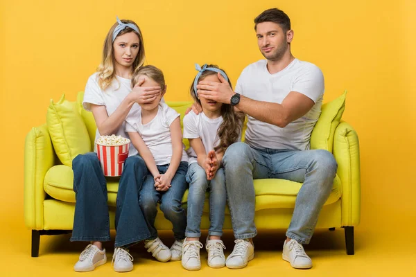 Skeptical parents closing eyes to kids while watching movie on sofa with popcorn bucket on yellow — Stock Photo
