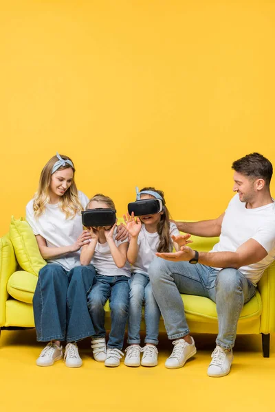 Parents joyeux avec des enfants dans des casques de réalité virtuelle assis sur le canapé sur jaune — Photo de stock