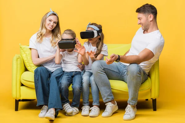 Parents heureux avec des enfants dans des casques de réalité virtuelle assis sur le canapé sur jaune — Photo de stock