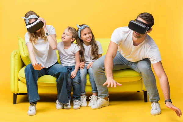 Laughing children sitting on sofa with parents in virtual reality headsets on yellow — Stock Photo