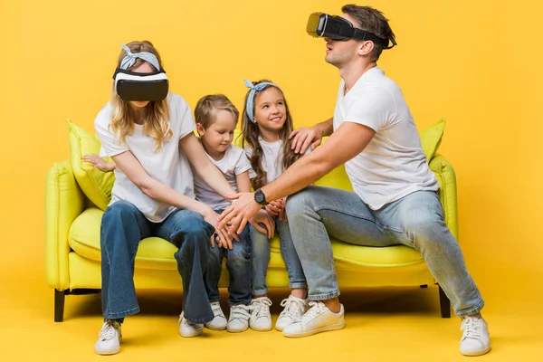 Happy children sitting on sofa with parents in virtual reality headsets on yellow — Stock Photo