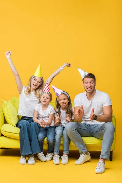 Glückliche Familie in Geburtstagskappen applaudiert und zeigt Daumen hoch auf Sofa auf gelb — Stockfoto