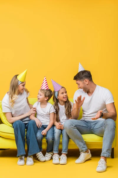 Cheerful family in birthday party caps applauding on sofa on yellow — Stock Photo