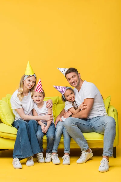 Cheerful family in birthday party caps hugging on sofa on yellow — Stock Photo