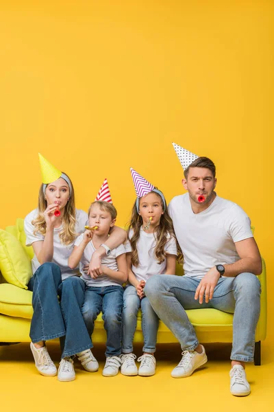 Padres y niños felices en gorras de fiesta de cumpleaños con sopladores en el sofá en amarillo - foto de stock