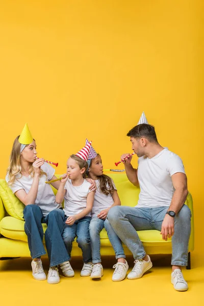 Fröhliche Familie in Geburtstagskappen mit Gebläse auf gelbem Sofa — Stockfoto