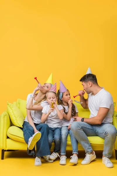 Happy parents and children in birthday party caps with blowers on sofa on yellow — Stock Photo