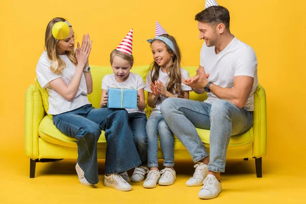 Famiglia allegra in berretti di festa di compleanno, figlio che tiene la scatola regalo sul divano su giallo — Foto stock