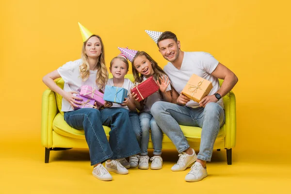 Cheerful family in birthday party caps holding gift boxes on sofa on yellow — Stock Photo