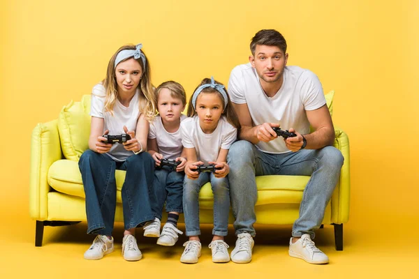 KIEW, UKRAINE - 4. MÄRZ 2020: Konzentrierte Familie spielt Videospiel mit Joysticks, während sie auf dem Sofa auf gelb sitzt — Stockfoto