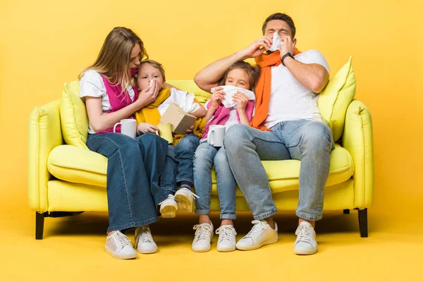 Ill family with kids holding napkins and having runny noses while sitting on sofa on yellow — Stock Photo