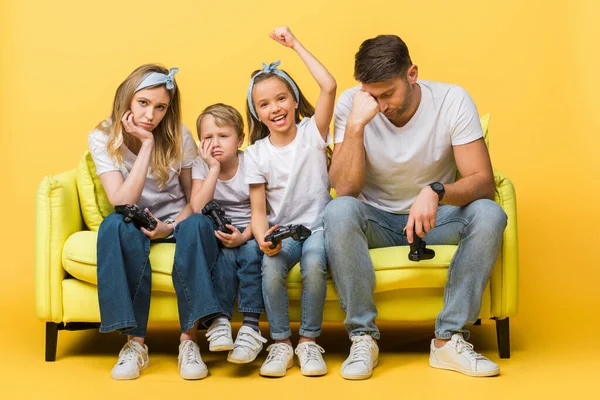 KYIV, UKRAINE - MARCH 4, 2020: upset family with joysticks sitting on sofa with happy daughter win in video game on yellow — Stock Photo