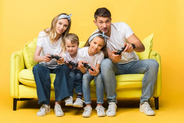 KYIV, UKRAINE - MARCH 4, 2020: cheerful family playing video game with joysticks while sitting on sofa on yellow — Stock Photo