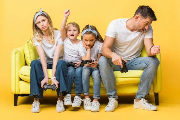 KIEW, UKRAINE - 4. MÄRZ 2020: traurige Familie mit Steuerknüppeln auf Sofa sitzend mit aufgeregtem Sohn gewinnt Videospiel auf gelb — Stockfoto