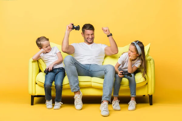 KYIV, UCRANIA - 4 de marzo de 2020: padre y niños emocionados jugando videojuegos con joysticks en el sofá en amarillo - foto de stock