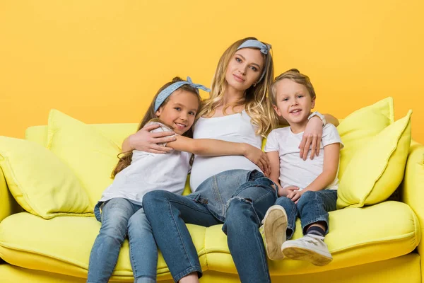 Happy pregnant mother hugging with kids on sofa isolated on yellow — Stock Photo