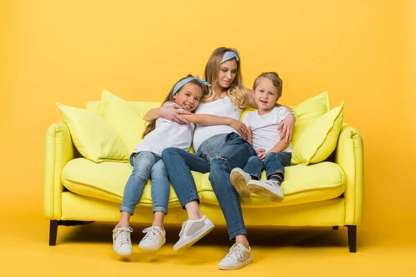 Mãe grávida feliz abraçando com irmãos no sofá no amarelo — Fotografia de Stock