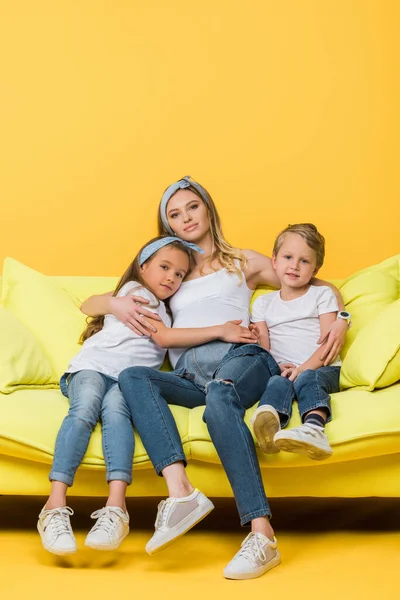 Pregnant mother hugging with daughter and son on sofa on yellow — Stock Photo