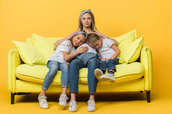 Embarazada madre abrazo con sonriente hija e hijo en sofá en amarillo - foto de stock