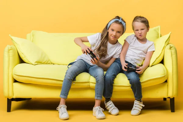 KYIV, UCRANIA - 4 de marzo de 2020: niños emocionados jugando videojuegos con joysticks en el sofá en amarillo - foto de stock
