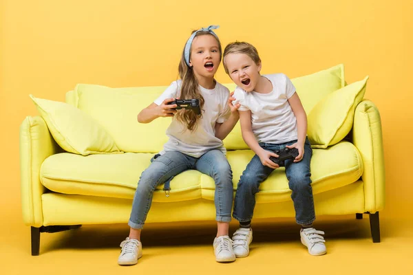 KYIV, UKRAINE - MARCH 4, 2020: emotional siblings screaming and playing video game with joysticks while sitting on sofa on yellow — Stock Photo