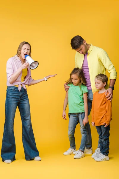 Angry mother yelling into megaphone on husband and sad children on yellow — Stock Photo