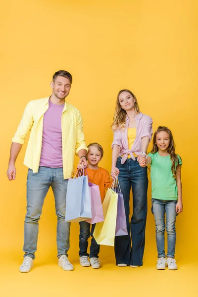 Padres alegres tomados de la mano con los niños mientras sostienen bolsas de compras en amarillo - foto de stock