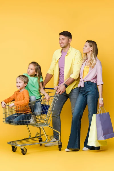 Pais felizes com sacos de compras e crianças sentadas no carrinho de compras em amarelo — Fotografia de Stock
