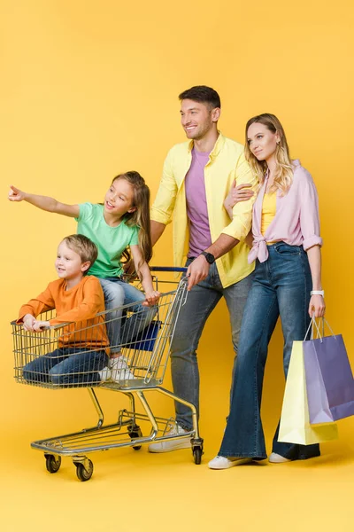 Padres con bolsas de compras y niños haciendo gestos y sentados en el carrito de la compra en amarillo - foto de stock