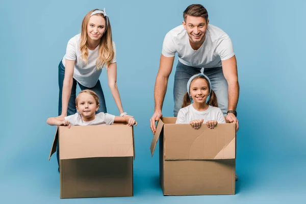 Smiling parents with children in cardboard boxes for relocation on blue — Stock Photo