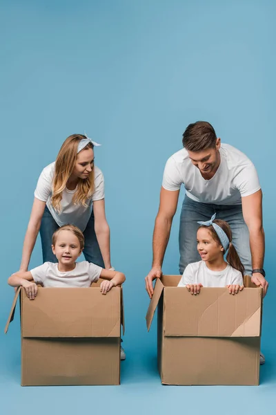 Happy parents with cheerful kids in cardboard boxes for relocation on blue — Stock Photo