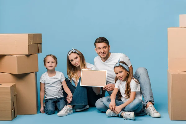 Happy parents and kids holding frame and sitting on blue with cardboard boxes for relocation — Stock Photo