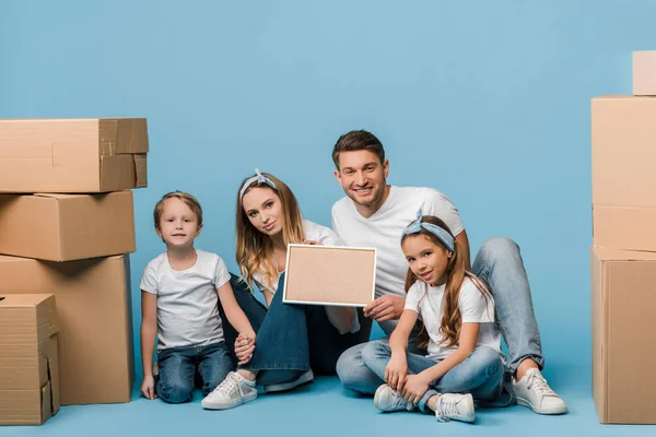 Genitori felici e bambini che tengono la cornice e seduti su blu con scatole di cartone per il trasferimento — Foto stock