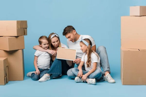 Positive parents and kids holding frame and sitting on blue with cardboard boxes for relocation — Stock Photo