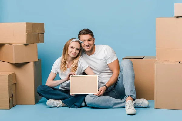 Happy couple holding empty frame while sitting on blue with cardboard boxes for relocation — Stock Photo