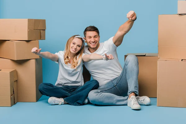 Aufgeregtes Paar zeigt Daumen hoch, während es mit Pappkartons für den Umzug auf blauem Grund sitzt — Stockfoto